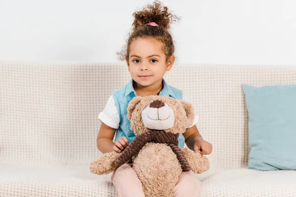Adorable enfant afro-américain assis sur le canapé avec ours en peluche et souriant à la caméra — Photo de stock