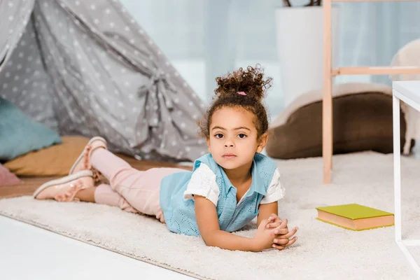 Adorable enfant afro-américain couché sur le tapis avec un livre et regardant la caméra — Photo de stock