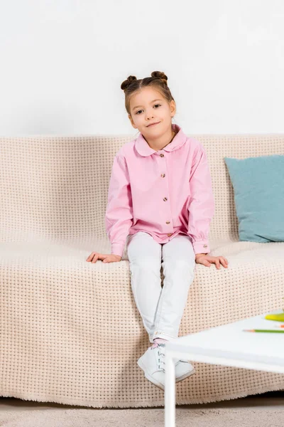 Ganzkörperansicht des entzückenden kleinen Kindes, das auf der Couch sitzt und in die Kamera lächelt — Stockfoto