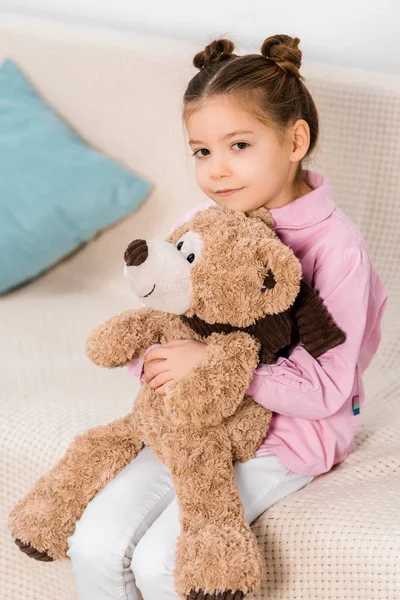 Чарівна дитина сидить на дивані з плюшевим ведмедем і посміхається на камеру — стокове фото