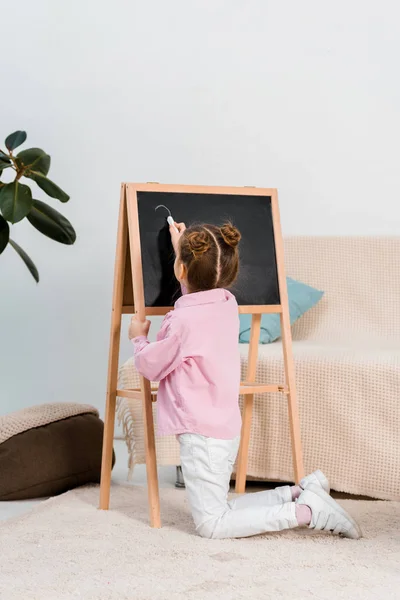 Rückansicht des niedlichen Kindes, das auf Teppich kniet und auf Kreidetafel schreibt — Stockfoto