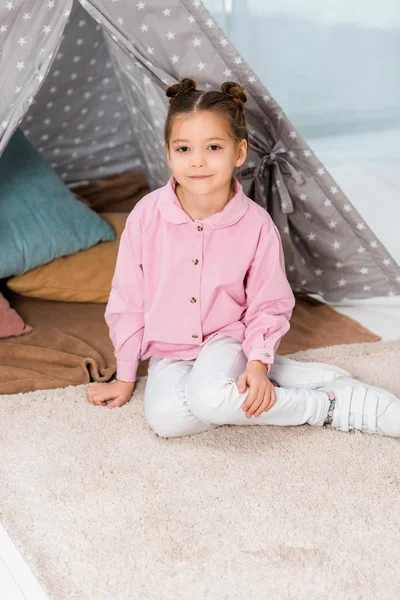 Beautiful happy child sitting on carpet near teepee and smiling at camera — Stock Photo