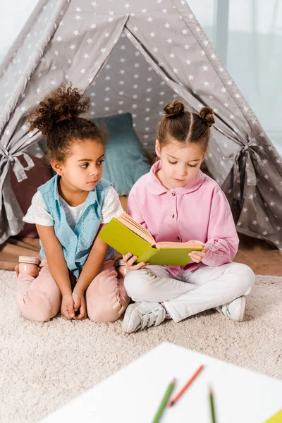 Adorable multiethnic kids sitting on carpet and reading book together — Stock Photo