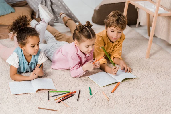 Vista de ángulo alto de adorables niños multiétnicos acostados en la alfombra y estudiando juntos - foto de stock