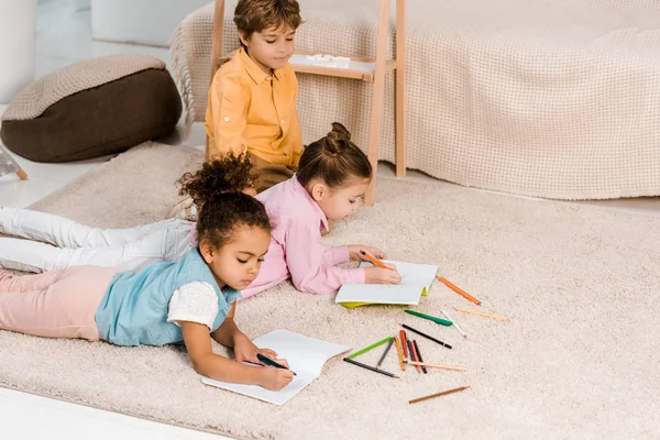 Vista de ángulo alto de hermosos niños multiétnicos acostados en la alfombra y estudiando juntos - foto de stock