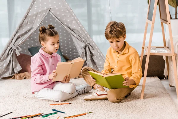 Hermosos niños enfocados sentados en la alfombra y leyendo libros juntos - foto de stock