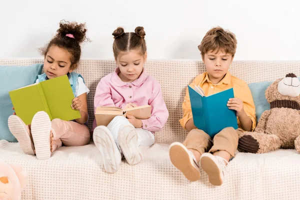 Entzückende multiethnische Kinder sitzen auf dem Sofa und lesen gemeinsam Bücher — Stockfoto