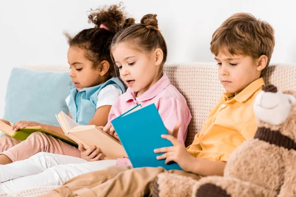 Beaux enfants multiethniques assis sur le canapé et lisant des livres ensemble — Photo de stock