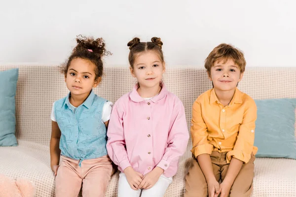 Adorable multiethnic children sitting on sofa and smiling at camera — Stock Photo