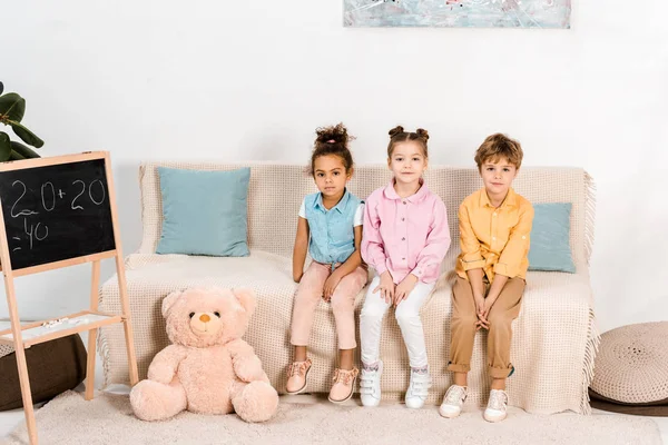 Beautiful multiethnic children sitting on couch and looking at camera — Stock Photo