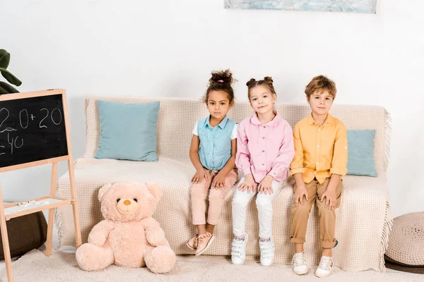 Entzückende kleine multiethnische Kinder, die auf der Couch sitzen und in die Kamera schauen — Stockfoto