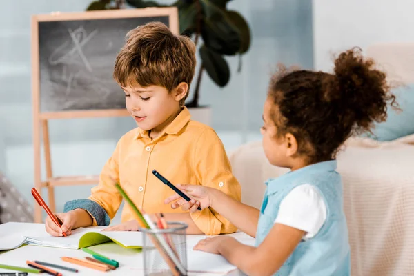 Niedliche multiethnische Kinder sitzen und schreiben mit Stiften zusammen — Stockfoto