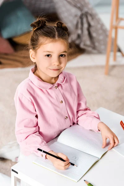 Criança adorável escrevendo com lápis e sorrindo para a câmera — Fotografia de Stock