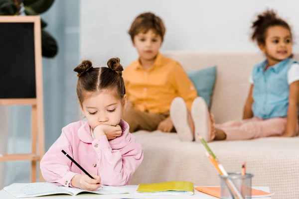 Adorable little child writing with pencil while friends sitting behind — Stock Photo