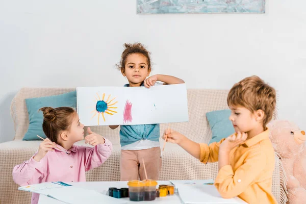Cute african american child showing picture while painting with friends — Stock Photo