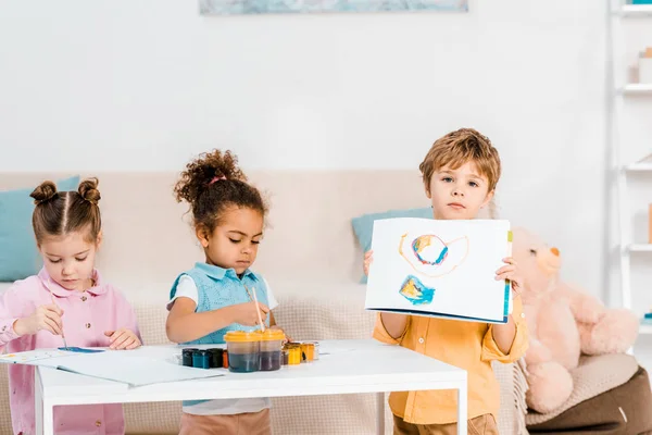 Cute little boy showing picture while painting with friends — Stock Photo