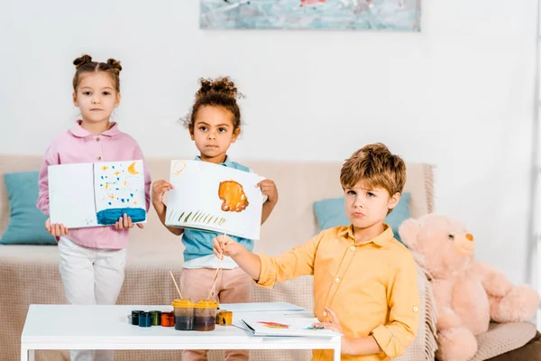Adorable multiethnic children holding pictures and looking at camera while painting together — Stock Photo
