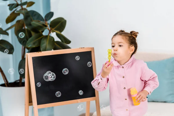 Adorable child standing near blackboard and blowing soap bubbles — Stock Photo