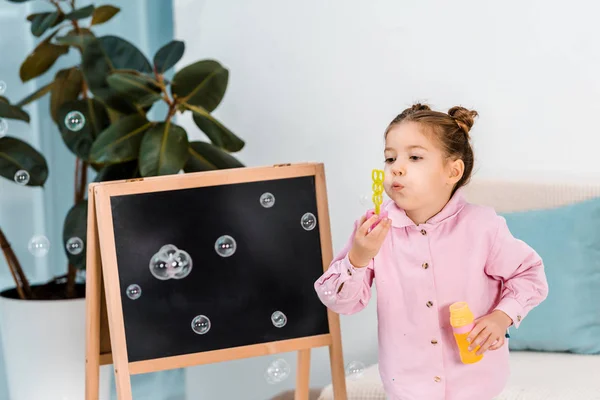 Criança adorável de pé perto de quadro-negro e soprando bolhas de sabão — Fotografia de Stock