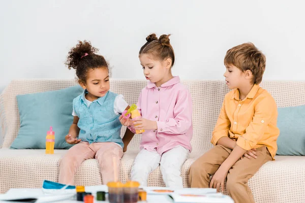 Adorables enfants multiethniques assis sur le canapé et jouant avec des bulles de savon — Photo de stock