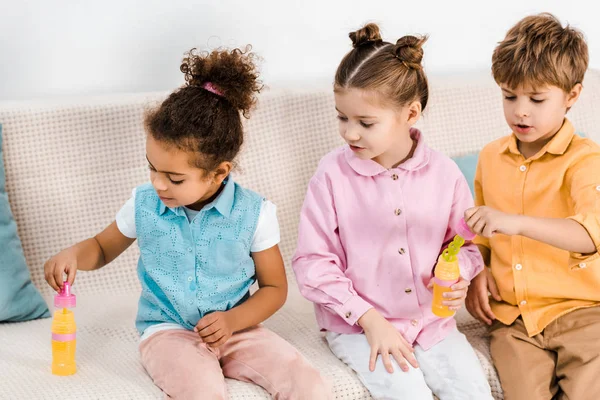 Adorable multiethnic children sitting on sofa and blowing soap bubbles — Stock Photo