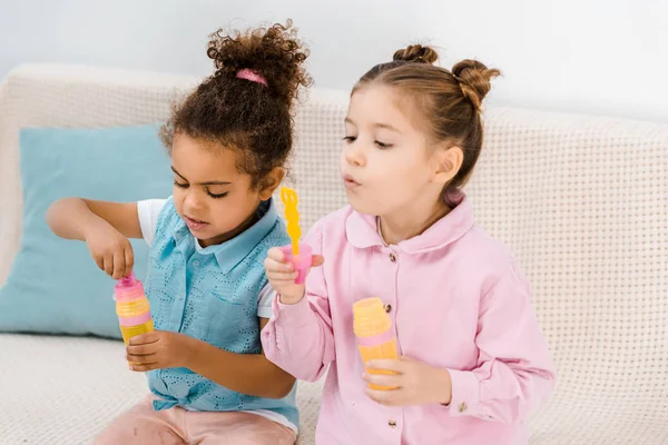 Bellissimi bambini multietnici seduti sul divano e che soffiano bolle di sapone — Foto stock