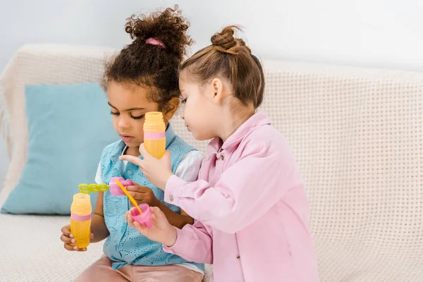 Adorable multiethnic children sitting on sofa and blowing soap bubbles — Stock Photo