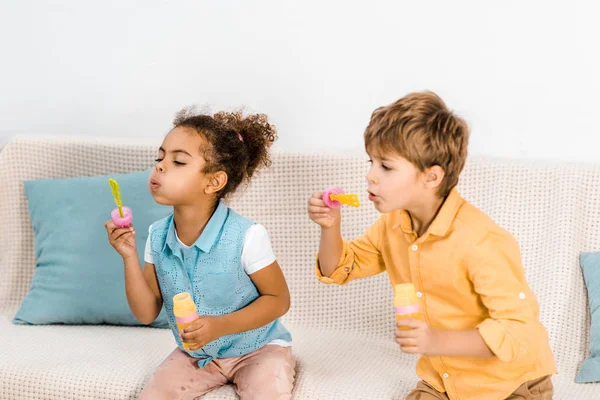 Adorables niños multiétnicos sentados en el sofá y soplando burbujas de jabón - foto de stock
