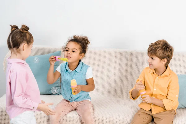 Adorable multiethnic kids sitting on sofa and blowing soap bubbles — Stock Photo