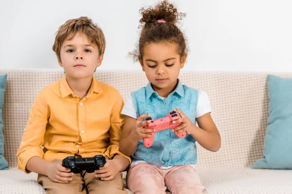 Belas crianças multiétnicas sentadas no sofá e brincando com joysticks — Fotografia de Stock