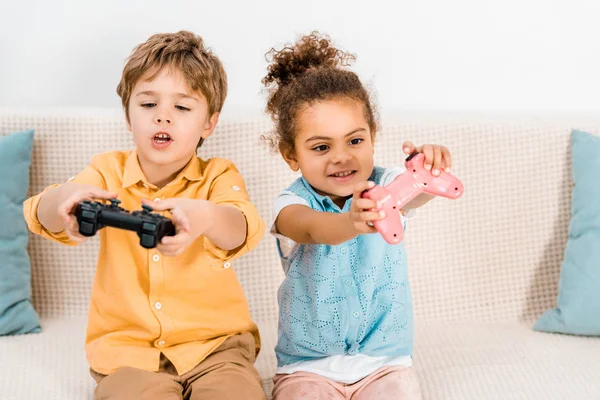 Милые эмоциональные мультиэтнические дети сидят на диване и играют в видеоигры с джойстиками — стоковое фото