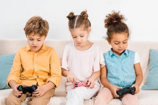 Adoráveis crianças multiétnicas sentadas no sofá e brincando com joysticks — Fotografia de Stock