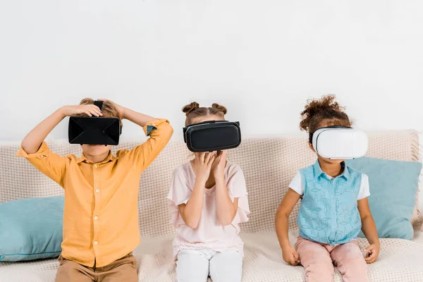 Adoráveis crianças multiétnicas sentadas no sofá e usando fones de ouvido realidade virtual — Fotografia de Stock