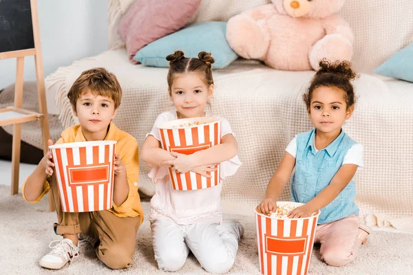 Adorables niños multiétnicos sosteniendo cajas con palomitas de maíz y mirando a la cámara - foto de stock