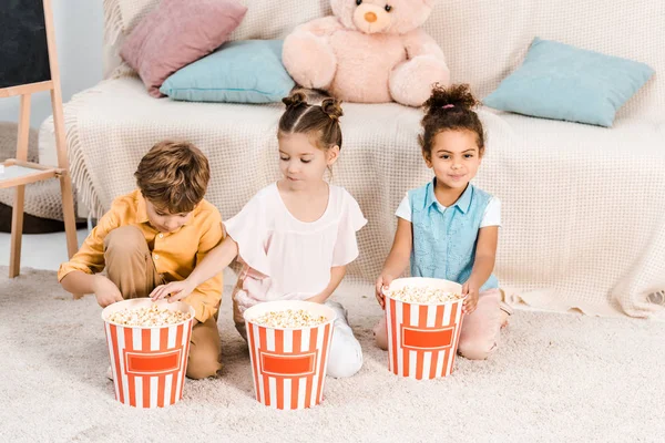 Vue grand angle de mignons enfants multiethniques assis sur le tapis et manger du maïs soufflé à partir de boîtes — Photo de stock