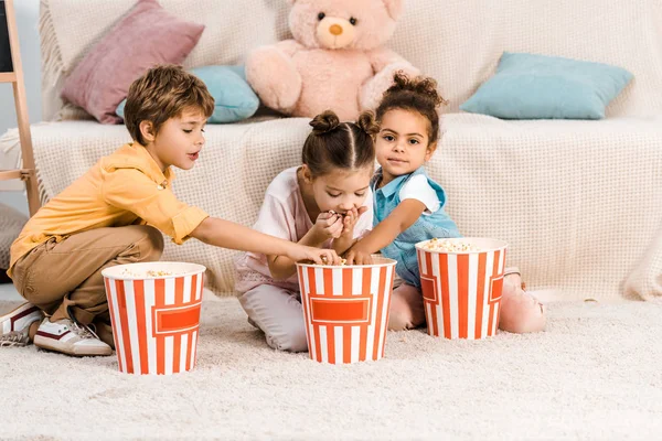 Милі багатоетнічні діти сидять на килимі і їдять попкорн — стокове фото