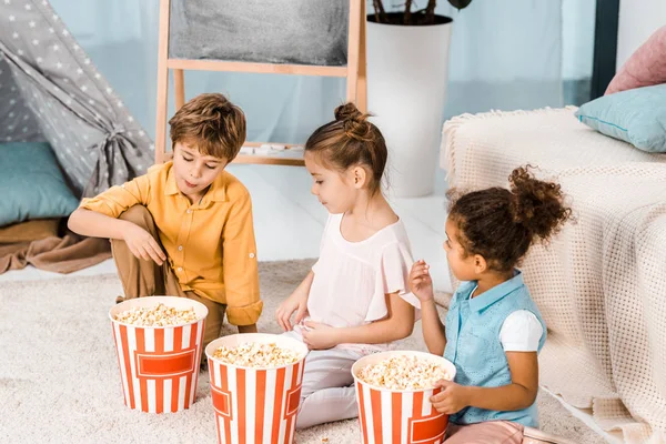 Cute little children sitting on carpet and eating popcorn — Stock Photo