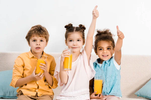 Adorables niños multiétnicos sosteniendo vasos de jugo y mostrando los pulgares hacia arriba - foto de stock