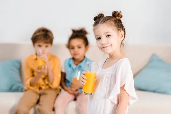 Adorable niño sosteniendo un vaso de jugo y sonriendo a la cámara mientras pequeños amigos bebiendo jugo detrás - foto de stock