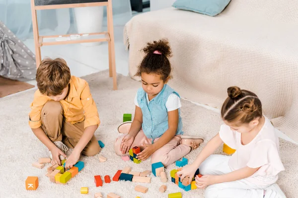 Vista de ángulo alto de adorables niños multirraciales jugando con cubos de colores en la alfombra - foto de stock