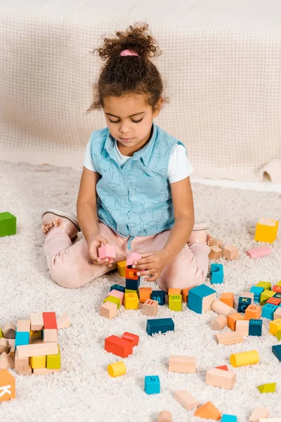 Vista de ángulo alto de niño afroamericano lindo sentado en la alfombra y jugando con bloques de madera de colores - foto de stock