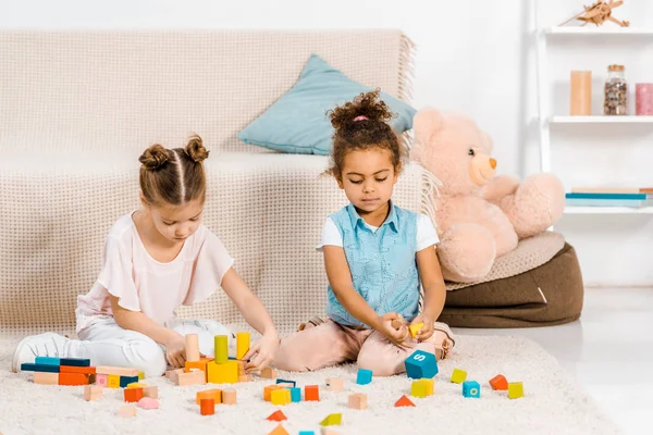 Hermosos niños multiétnicos pequeños sentados en la alfombra y jugando con cubos de colores - foto de stock