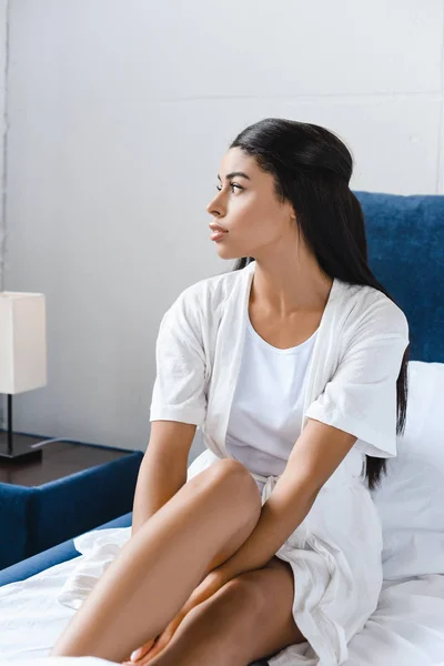 Attractive mixed race girl in white robe sitting on bed and looking away — Stock Photo