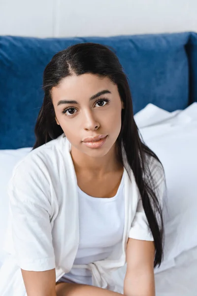 Retrato de hermosa chica de raza mixta en bata blanca sentado en la cama y mirando a la cámara en la mañana - foto de stock