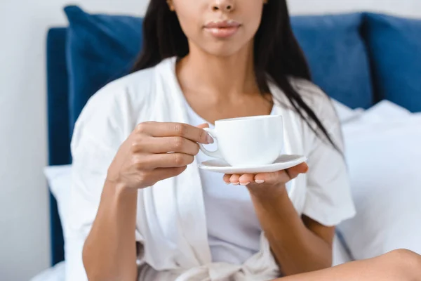 Imagem cortada de menina de raça mista em roupão branco segurando xícara de café na cama de manhã — Fotografia de Stock