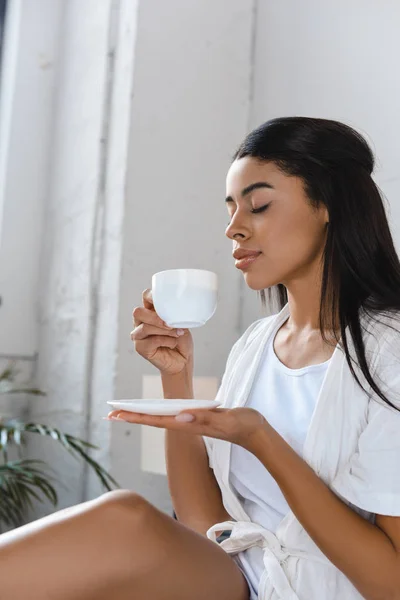 Vista laterale di bella ragazza razza mista in accappatoio bianco annusare caffè aromatico a letto al mattino — Foto stock