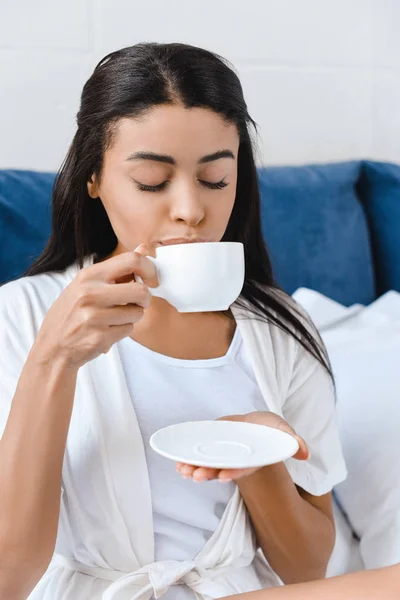 Приваблива змішана дівчина в білому халаті п'є каву в ліжку вранці з закритими очима — стокове фото