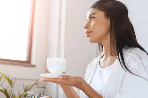 Vista lateral de hermosa chica de raza mixta en túnica blanca sosteniendo taza de café en la mañana en el dormitorio con luz solar - foto de stock