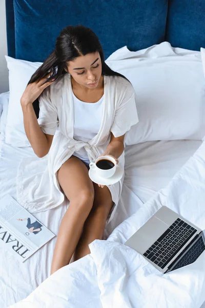 Vista de ángulo alto de hermosa chica de raza mixta en túnica blanca sosteniendo taza de café en la cama por la mañana y mirando el ordenador portátil - foto de stock