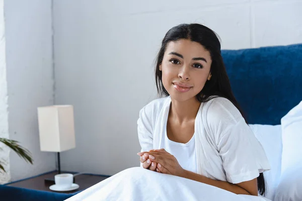 Hermosa chica de raza mixta en bata blanca sentado en la cama y mirando a la cámara en la mañana - foto de stock